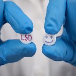 Gloved hands holding two pills, one labeled LSD and one with a smiley face