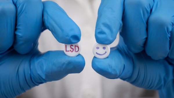 Gloved hands holding two pills, one labeled LSD and one with a smiley face